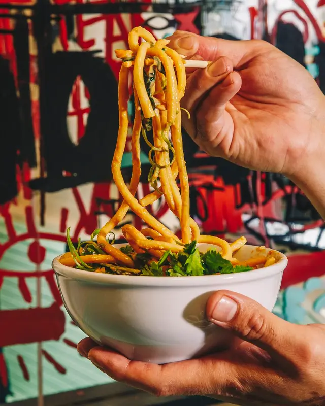 Handheld bowl of saucy udon with chopsticks and graffiti mural background
