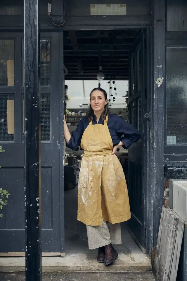 Artisan in doorway of urban ceramics studio wearing stained mustard apron