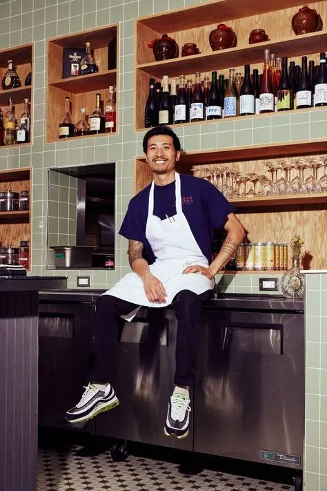 Smiling chef in white apron seated on stainless under-counter fridge at tiled bar