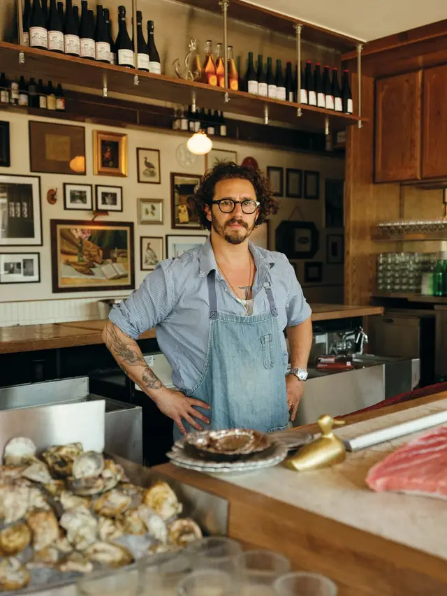 Oyster bar chef in denim apron at wood counter with oysters and wine