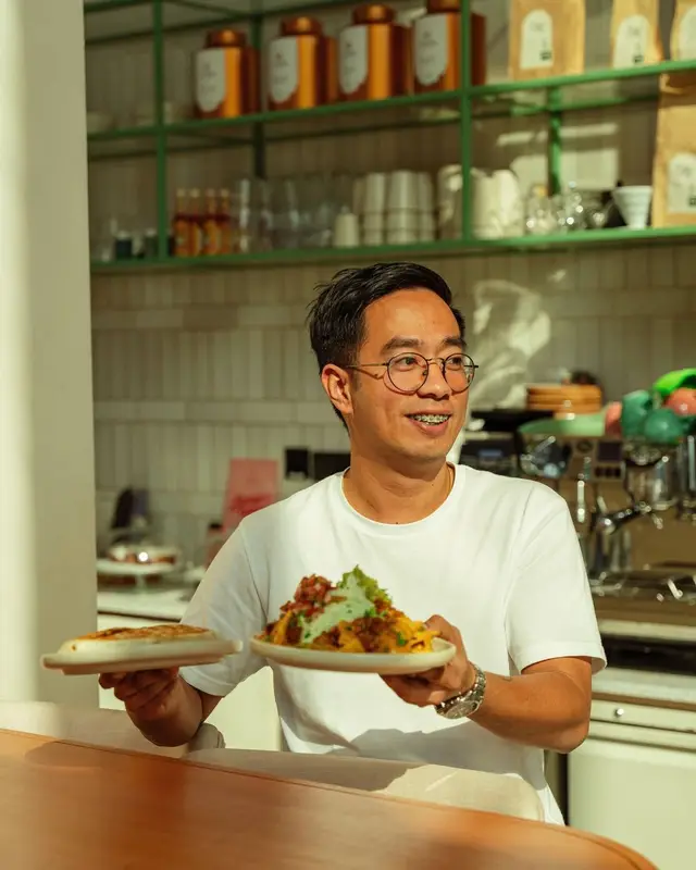 Sunlit café server carrying two plates of brunch at green-shelved counter