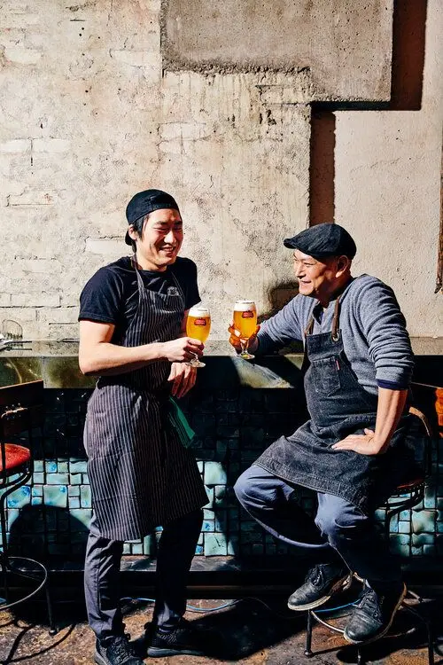 Two apron-clad bartenders clinking craft beer at a blue-tiled bar