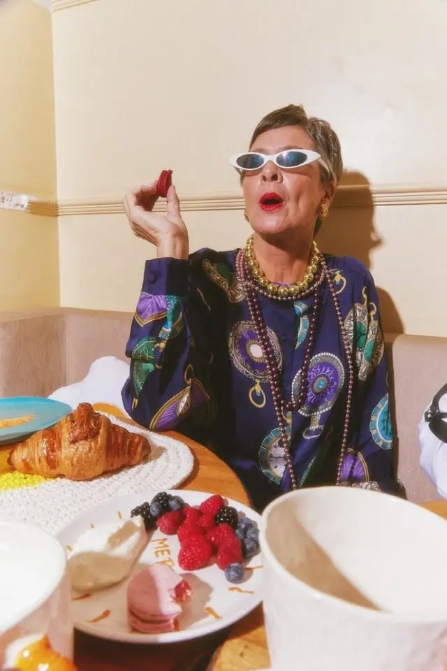Woman in patterned silk blouse with layered beaded necklaces eating pastries at a café