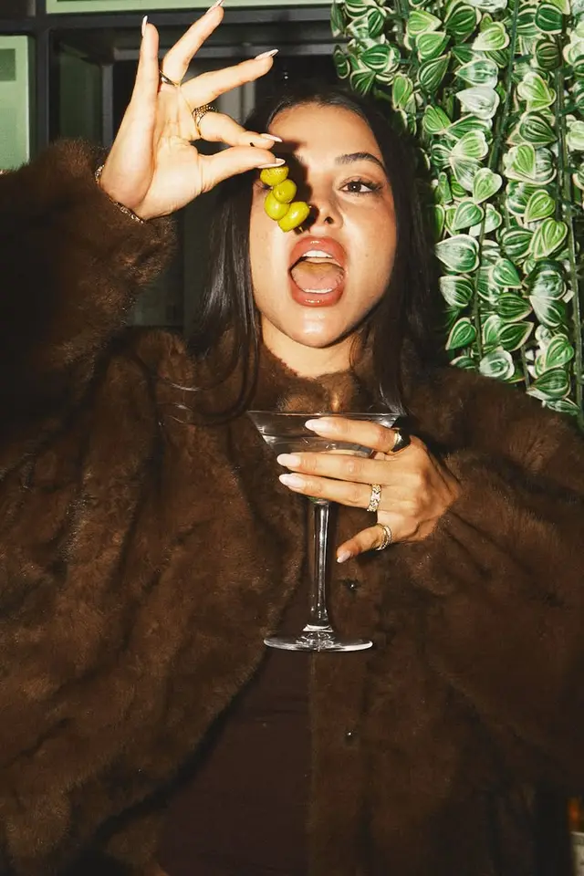 Flash-lit portrait: woman eating skewered olives over martini, brown fur coat