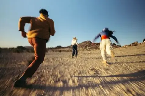 Three people running across a sunlit gravel desert, cinematic motion blur