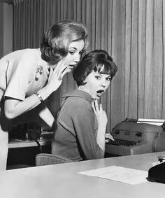 Mid-century office gossip — woman whispers to colleague at typewriter (black-and-white)