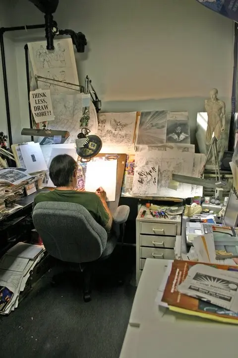 Illustrator at drafting table — studio with 'THINK LESS, DRAW MORE!!' sign