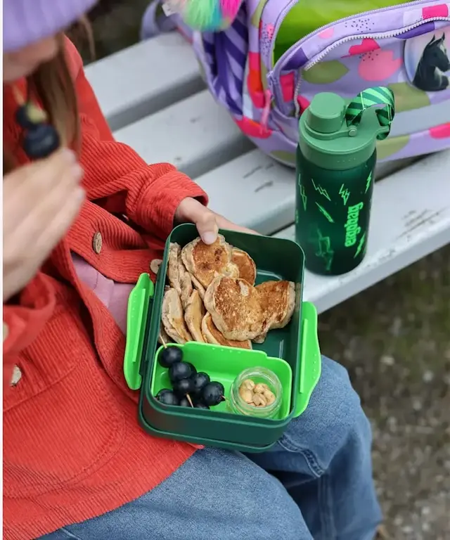 Green bento lunchbox with mini pancakes, grapes and nut cup on a bench
