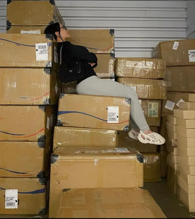 Person reclining on stacked shipping boxes in warehouse with black beanie and gray tights