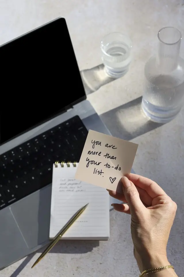Hand holding sticky note: "You are more than your to-do list" beside laptop, notebook, pen