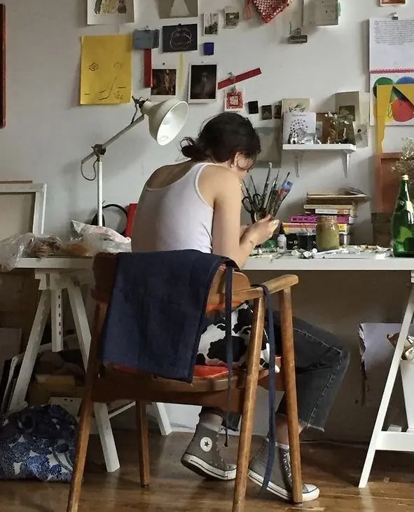 Artist at cluttered home studio desk with pinboard wall