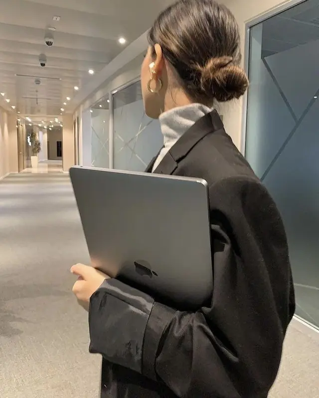 Person in black blazer holding space gray MacBook in modern office corridor