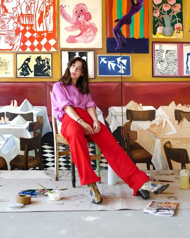 Person seated in front of large restaurant mural — red trousers, purple blouse