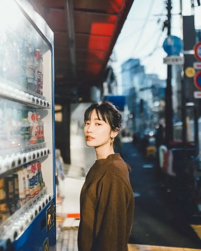 Woman in brown sweater beside blue beverage vending machine on city street