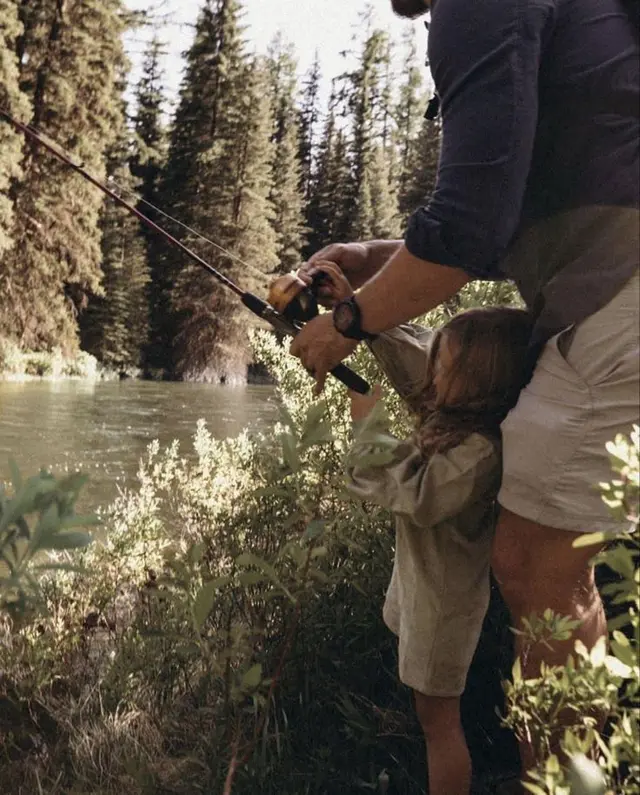 Adult guiding child to fish on pine-lined riverbank