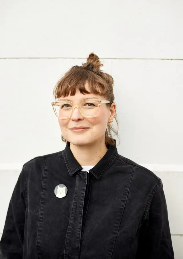Editorial headshot — clear acetate glasses, black denim jacket, neutral backdrop