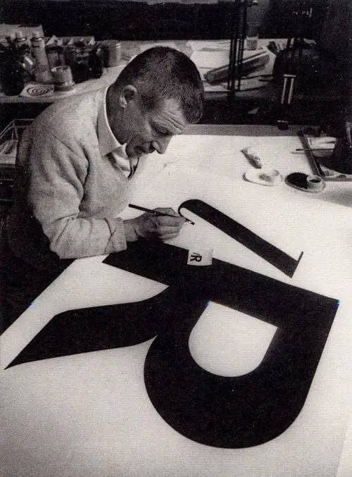 Black-and-white studio photo of designer painting oversized letter R on layout table