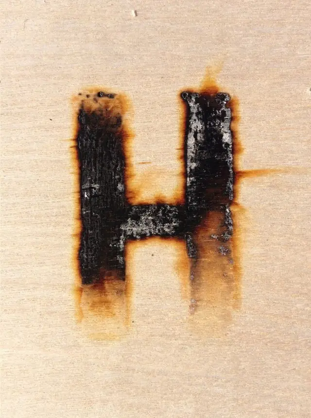 Burned letter "H" on light plywood — charred wood texture