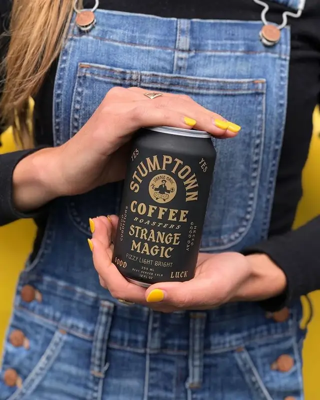 Person holding Stumptown Coffee 'Strange Magic' matte black can with denim overalls