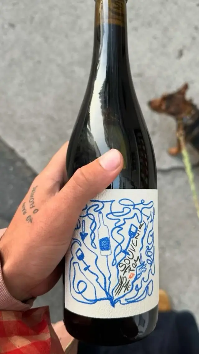 Hand-Held Red Wine Bottle with Blue Doodle Label Reading 'SOUVENIR FROM'