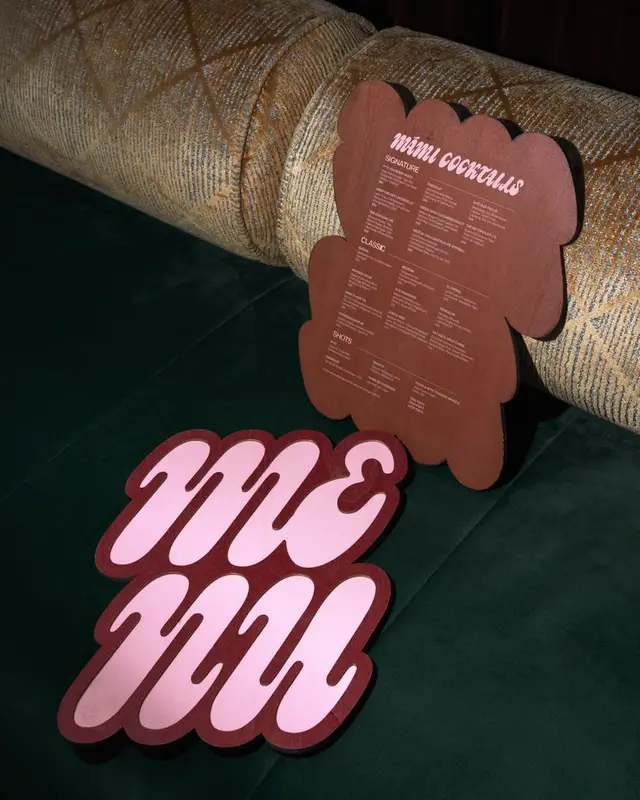 Die-cut maroon cocktail menu with pale-pink 'ME' plaque on green velvet bench