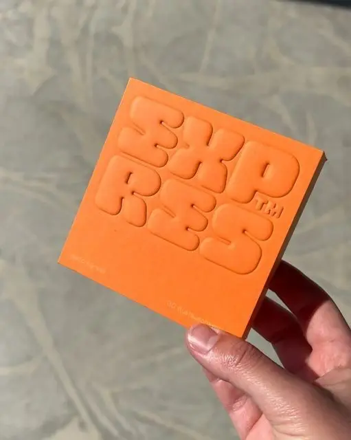 Hand holding small orange embossed square with bold rounded lettering