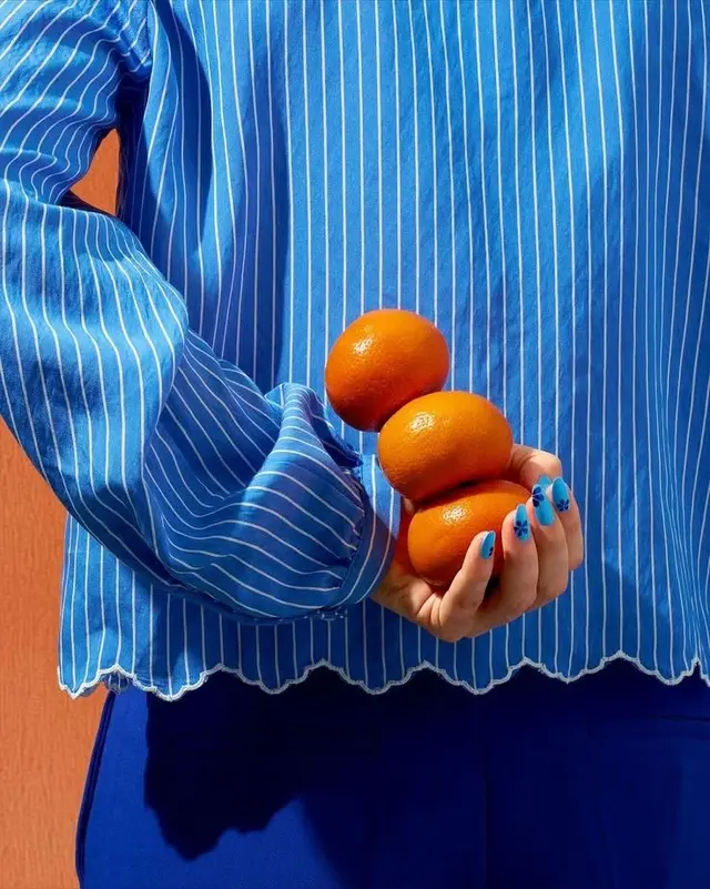 Cobalt pinstripe blouse holding three mandarins — blue-orange editorial still