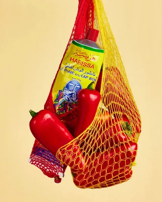 Harissa tube and red peppers in yellow mesh produce bag — product still life