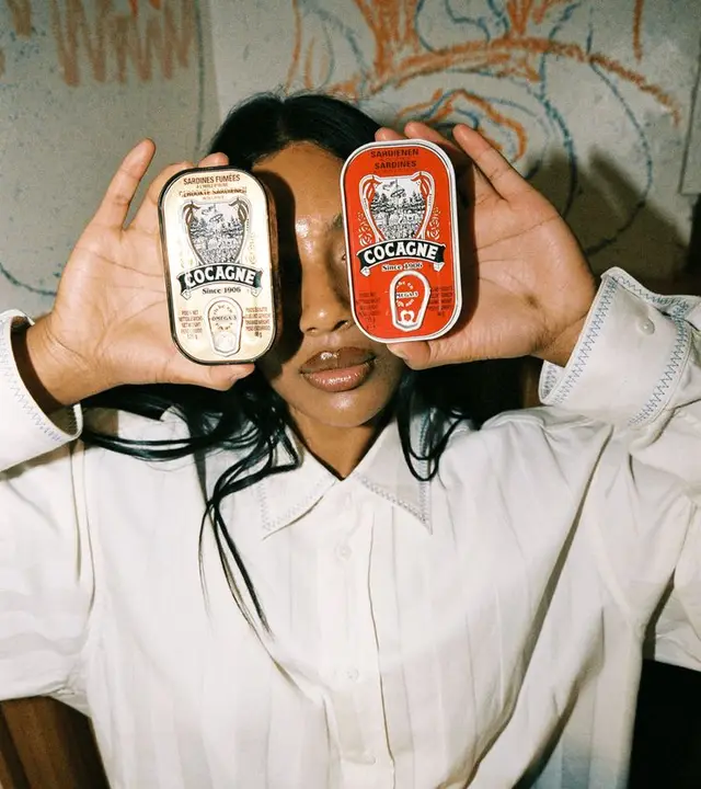 Editorial portrait — model holding Cocagne sardine tins (red and cream)
