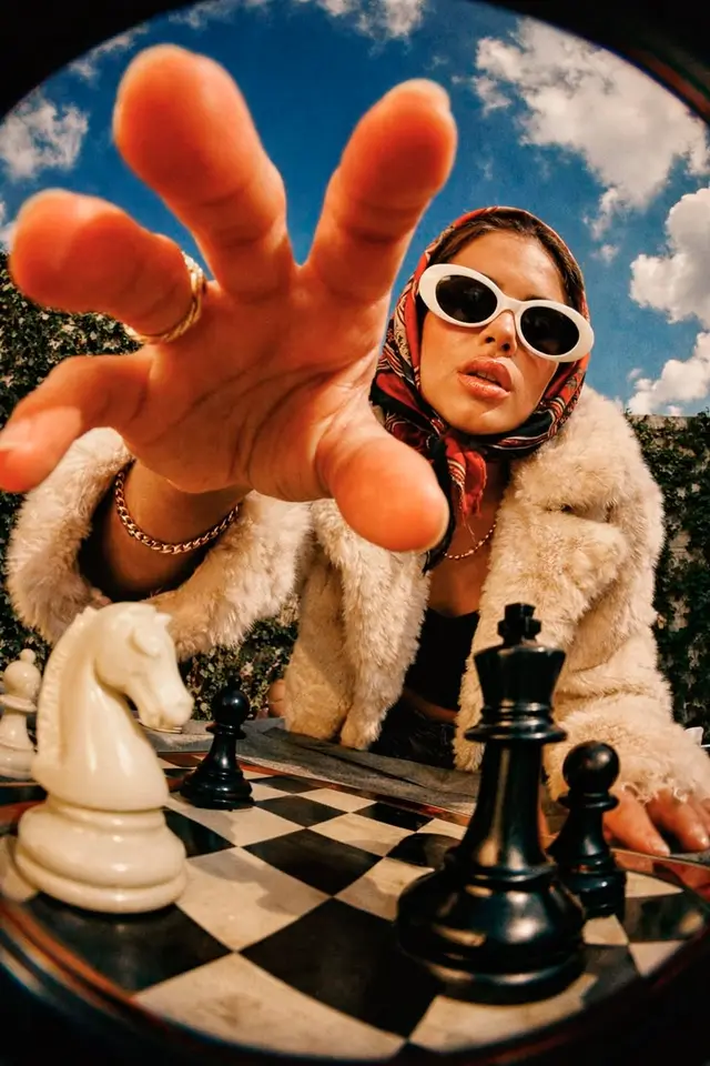 Low-angle fisheye portrait: woman in faux fur and headscarf reaching over chessboard