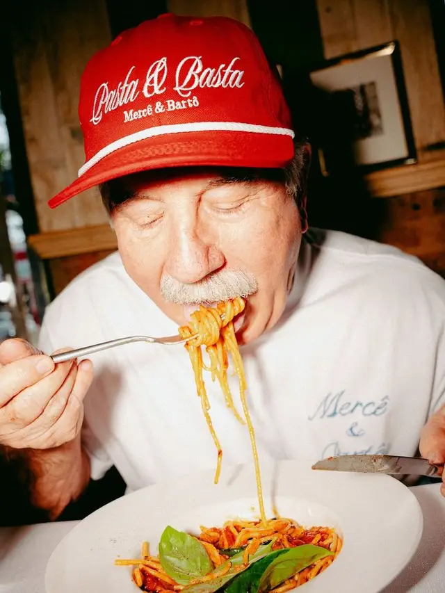 Man in red 'Pasta & Basta' cap eating spaghetti from a white bowl