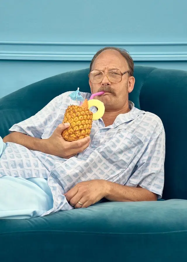 Man lounging on teal velvet sofa sipping pineapple cocktail
