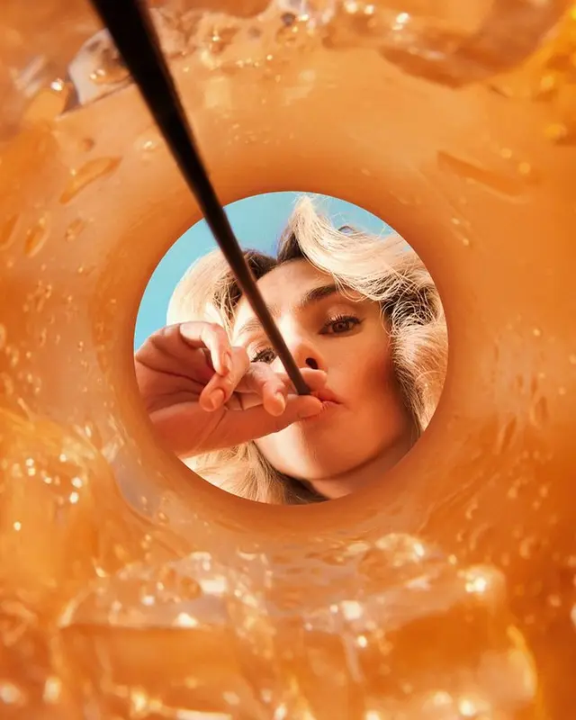 Inside-cup low-angle portrait — sipping iced orange drink through black straw
