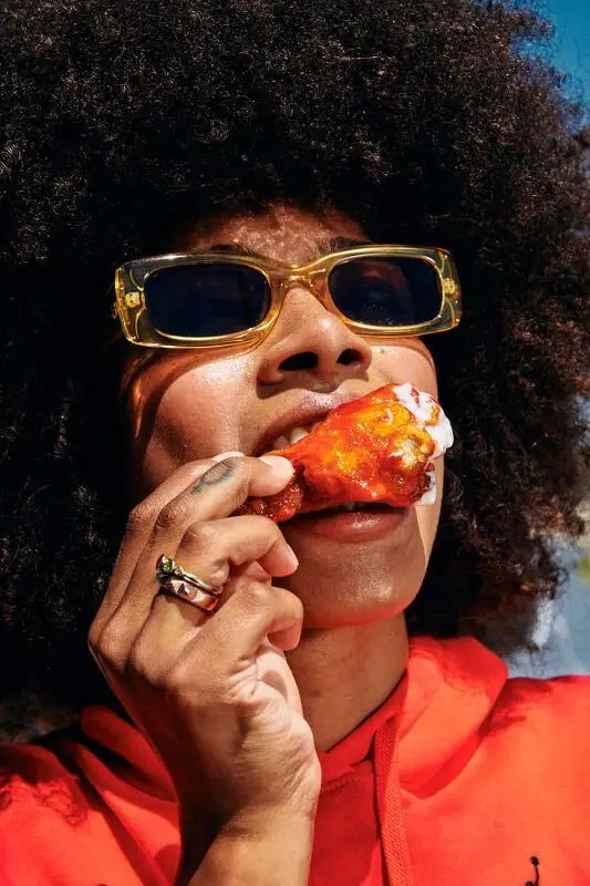 Close-up portrait of person eating sauced chicken wing in orange hoodie
