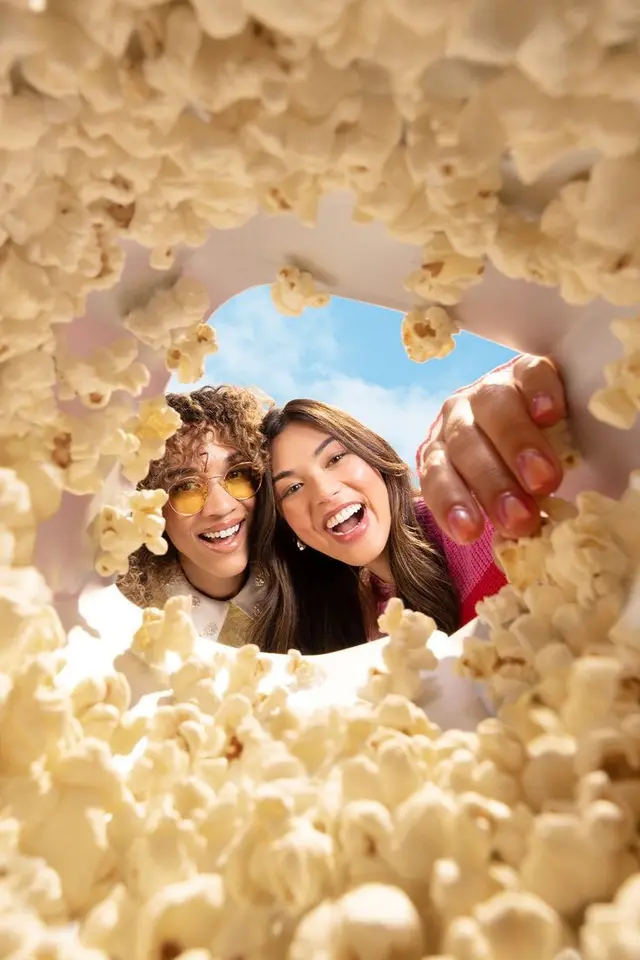 POV from inside a popcorn tub — two friends reaching in and laughing
