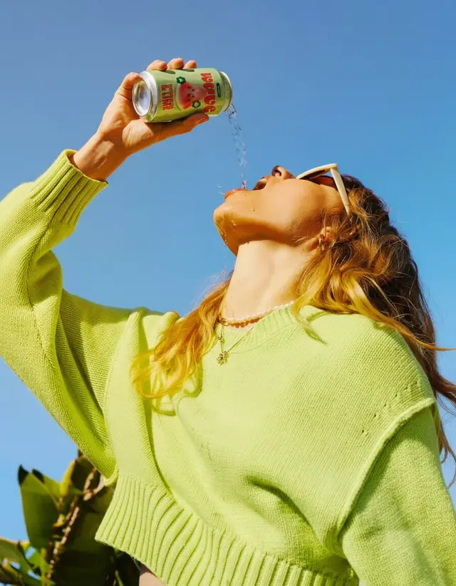 Person in lime knit sweater pouring drink from green can against blue sky