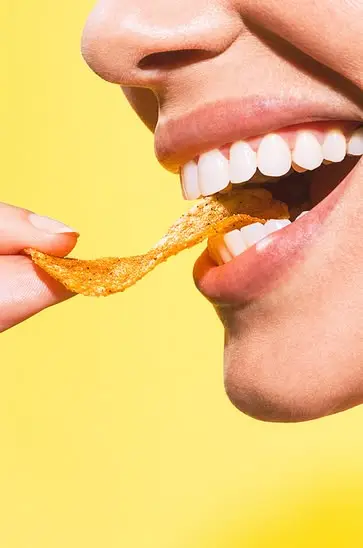 Close-up of mouth biting seasoned potato chip on bright yellow background