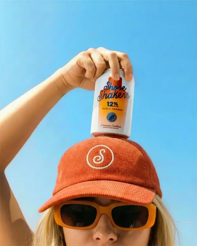 Shore Shaker blood orange canned cocktail held above orange corduroy cap