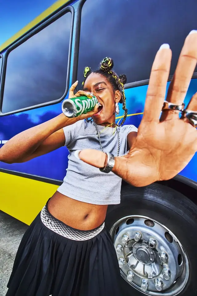 Close-up: person drinking Sprite in front of blue-and-yellow bus