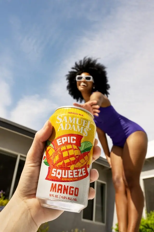Samuel Adams Epic Squeeze Mango can held in foreground with woman in purple swimsuit, low‑