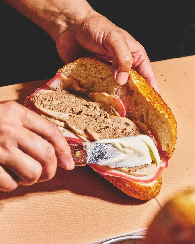 Crusty baguette with pâté and cured ham being spread with butter