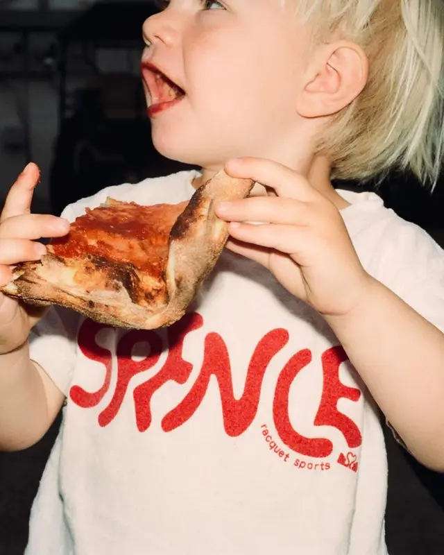 Candid close-up of toddler eating pizza in white 'SPENCE' racquet sports T-shirt