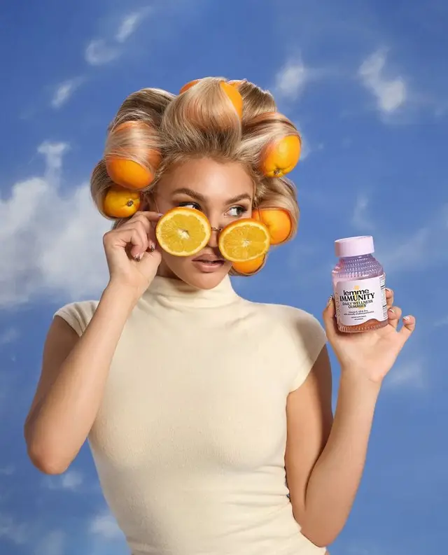 Woman with oranges-as-hair-rollers and orange-slice sunglasses holding a bottle labeled 'L