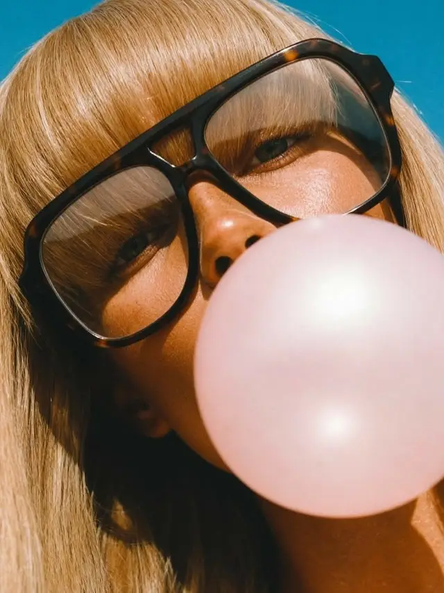Blonde close-up blowing pink bubble in oversized tortoiseshell glasses