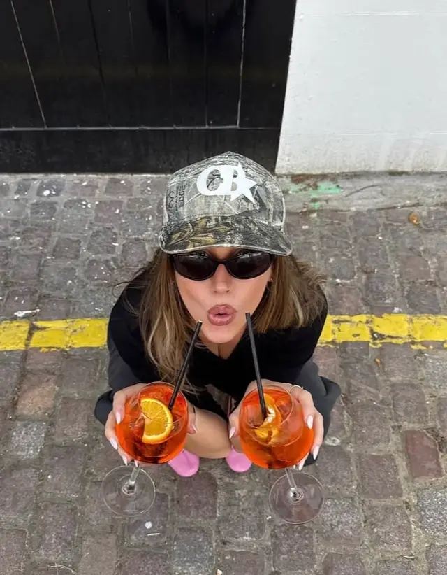 Top-down: person holding two orange spritz cocktails on cobblestone curb