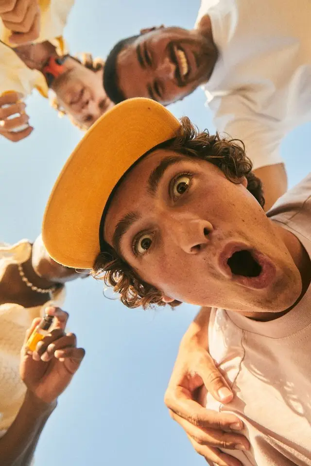 Low-angle candid of four friends leaning over camera — yellow baseball cap