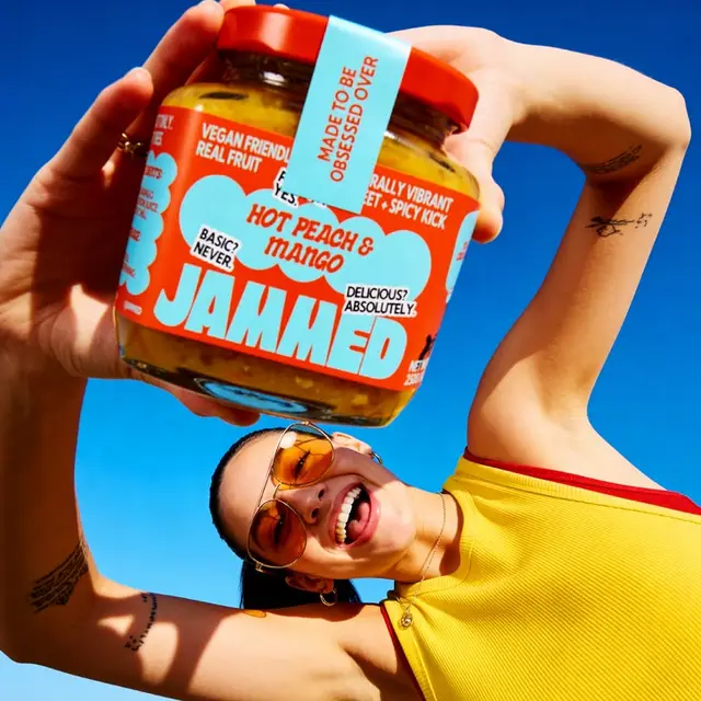 Person holding 'JAMMED' Hot Peach & Mango jam jar against bright blue sky