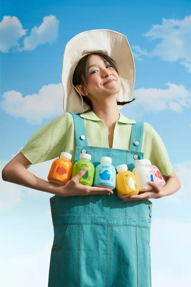 Smiling woman in teal overalls holding colorful bottled drinks against blue sky