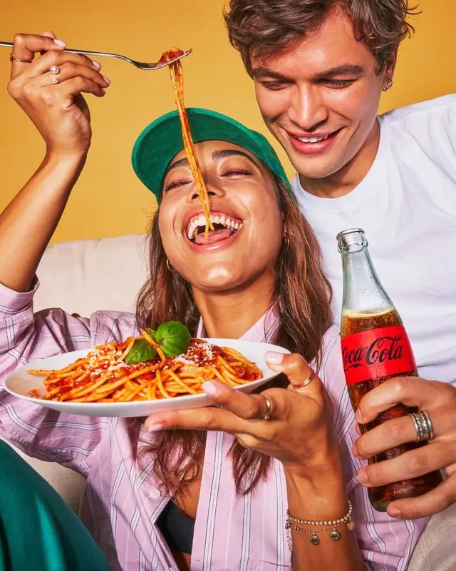 Couple sharing spaghetti with Coca‑Cola glass bottle on yellow backdrop