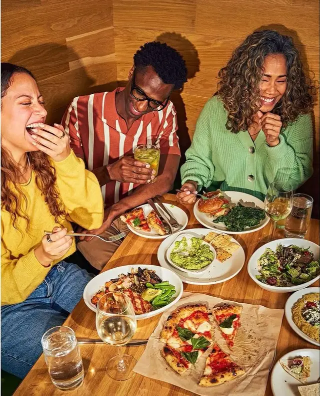 Friends sharing margherita pizza, bowls and drinks in wood-paneled booth